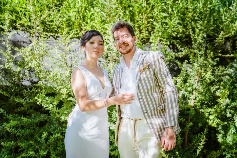 Hombre con chaqueta a rayas y mujer con vestido blanco posan juntos en un sendero rodeado de vegetación.