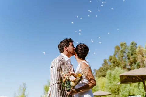 Hombre con chaqueta a rayas y mujer con vestido blanco se besan mientras varios globos blancos ascienden en el cielo.