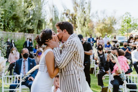Hombre con chaqueta a rayas y mujer con vestido blanco se besan en un jardín, con varias personas sentadas alrededor.