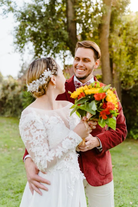 Hombre con chaqueta roja abraza por detrás a una mujer con vestido blanco, quien sostiene un ramo de girasoles y flores naranjas.