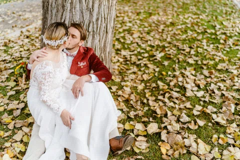 Hombre con chaqueta roja besa a una mujer con vestido blanco mientras ambos están sentados al pie de un árbol.