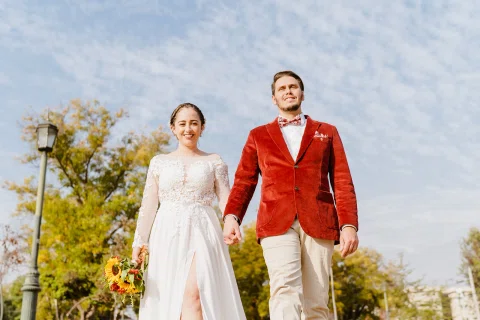 Hombre con chaqueta roja y mujer con vestido blanco caminan hacia la cámara tomados de la mano; ella sostiene un ramo.