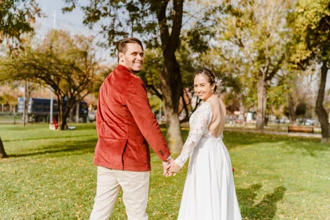 Hombre con chaqueta roja y mujer con vestido blanco caminan de la mano por el césped mientras miran hacia la cámara.