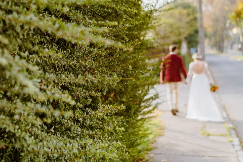 Hombre con chaqueta roja y mujer con vestido blanco caminan de espaldas por un sendero junto a un seto alto.