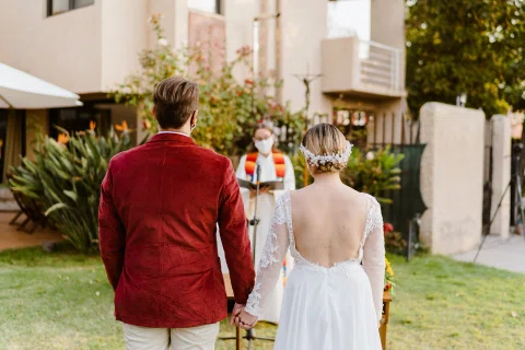 Hombre con chaqueta roja y mujer con vestido blanco están de pie y tomados de la mano, vistos de espaldas en un jardín.