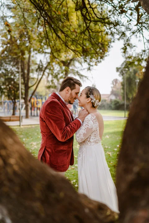 Hombre con chaqueta roja y mujer con vestido blanco permanecen frente a frente con los rostros próximos junto a un árbol.