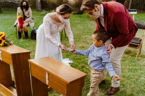 Hombre con chaqueta roja y mujer con vestido blanco se inclinan hacia un niño junto a dos muebles de madera en el jardín.