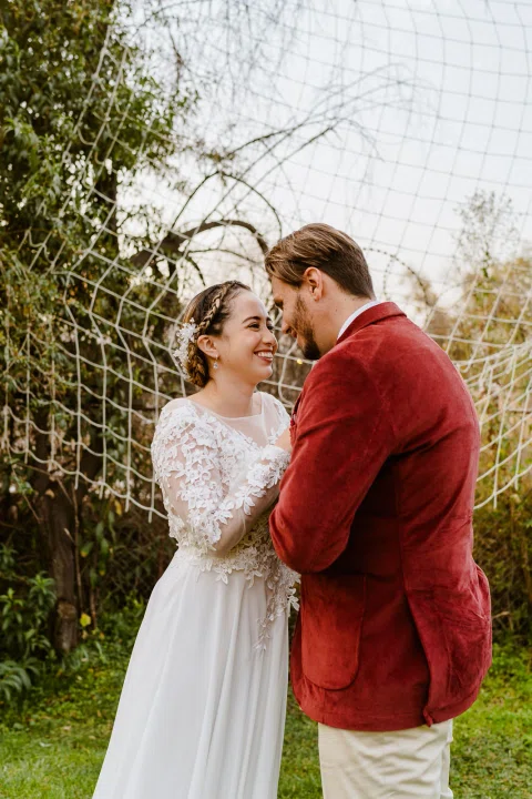 Hombre con chaqueta roja y mujer con vestido blanco posan juntos en un jardín.