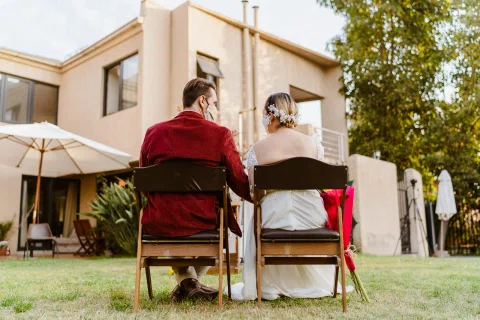 Hombre con chaqueta roja y mujer con vestido blanco están sentados de espaldas, con una casa y el jardín al fondo.