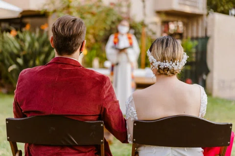 Hombre con chaqueta roja y mujer con vestido blanco están sentados de espaldas en un jardín, frente a una persona desenfocada.