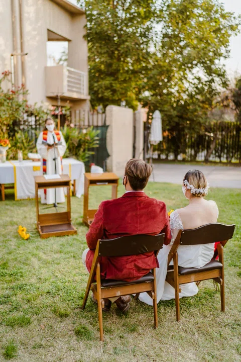 Hombre con chaqueta roja y mujer con vestido blanco están sentados de espaldas en un jardín con una casa al fondo.
