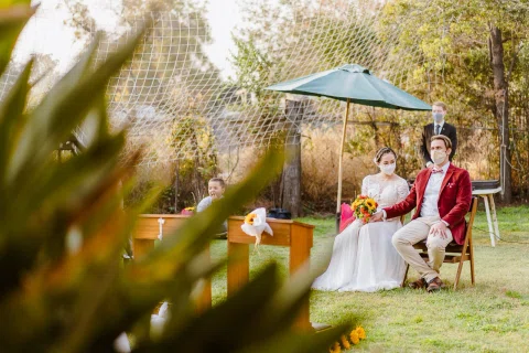 Hombre con chaqueta roja y mujer con vestido blanco están sentados juntos en un jardín; ella sostiene un ramo.