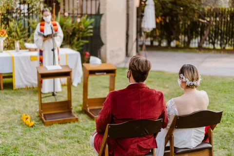 Vista amplia de un jardín con un hombre de chaqueta roja y una mujer de vestido blanco sentados de espaldas frente a una persona con túnica blanca.