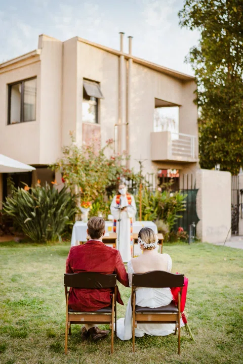 Vista vertical de un hombre con chaqueta roja y una mujer con vestido blanco sentados de espaldas en un jardín.