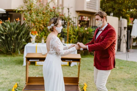Hombre con chaqueta roja y mujer con vestido blanco están de pie frente a frente y se toman de ambas manos en un jardín.