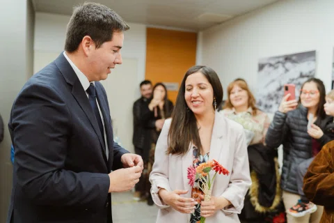Hombre con traje oscuro coloca un anillo en el dedo de una mujer que sostiene flores.