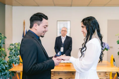 Hombre de traje negro coloca un anillo en la mano de una mujer con vestido blanco.