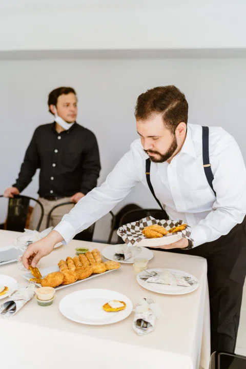 Hombre con tirantes coloca piezas de comida frita en un plato sobre una mesa mientras otra persona permanece detrás.