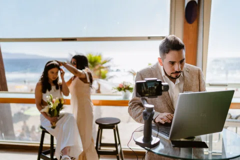Hombre trabaja frente a un computador junto a una cámara, mientras una mujer sentada con ramo y otra mujer aparecen junto al ventanal con vista al mar.