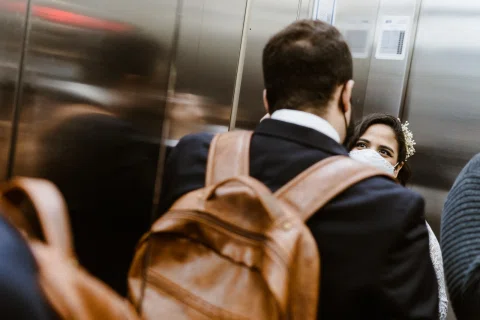 Hombre con mochila visto de espaldas y mujer con accesorio floral en el cabello dentro de un ascensor.