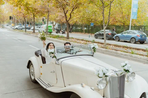 Hombre con lentes conduce un automóvil convertible blanco mientras una mujer con ramo ocupa el asiento del acompañante.