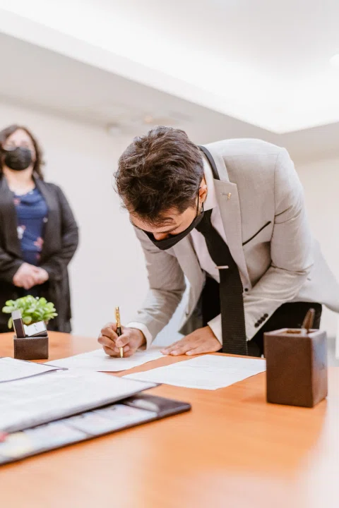 Hombre inclinado firmando un documento sobre una mesa.