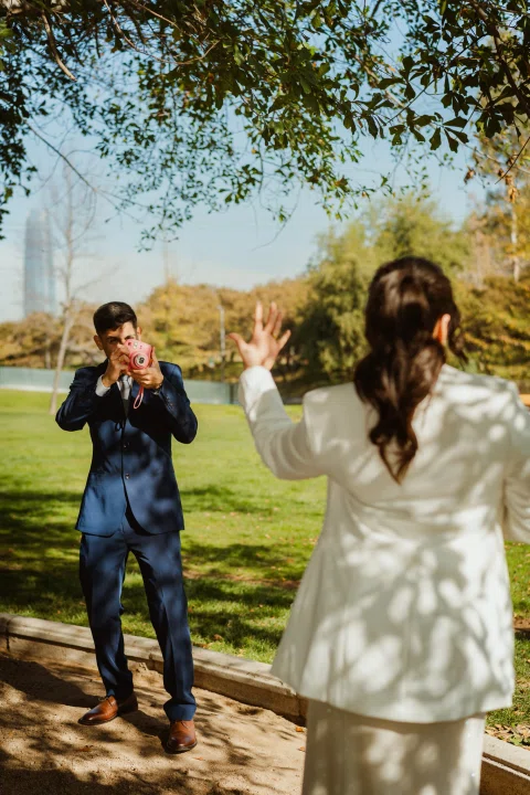 Hombre con traje azul fotografía con una cámara instantánea rosada a una mujer con traje claro vista de espaldas.