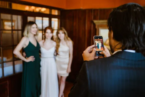 Hombre fotografía con un celular a tres mujeres que posan dentro de un salón.