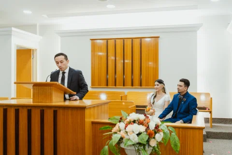 Hombre habla desde un atril de madera mientras una mujer y un hombre permanecen sentados al fondo.