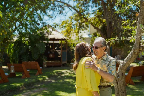 Hombre con lentes abraza a una mujer con blusa amarilla junto a un árbol en el jardín.