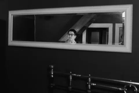 Hombre con lentes aparece reflejado en un espejo horizontal sobre una cama, en blanco y negro.