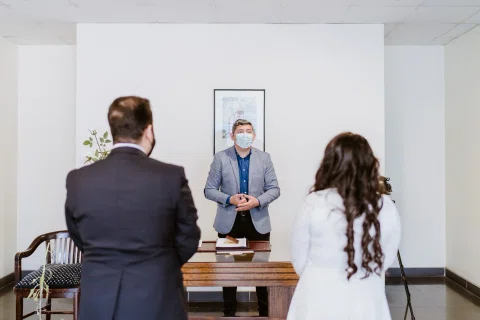 Hombre con mascarilla permanece detrás de una mesa frente a un hombre con traje oscuro y una mujer con vestido blanco, vistos de espaldas.