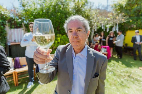 Hombre mayor sostiene una copa de vino hacia la cámara en el jardín.