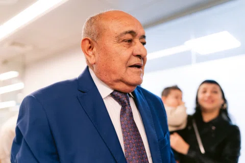 Hombre mayor con traje azul aparece en primer plano dentro de una sala luminosa.