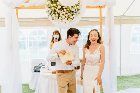 Hombre con camisa blanca muestra una libreta abierta junto a una mujer con vestido blanco que sonríe, bajo una decoración floral.