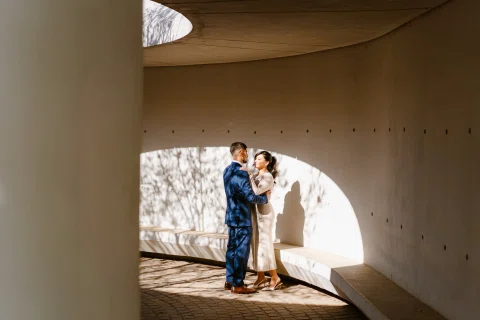 Hombre con traje azul y mujer con traje claro permanecen abrazados dentro de una estructura curva iluminada por luz lateral.