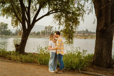 Hombre y mujer se abrazan bajo un árbol grande junto al agua.