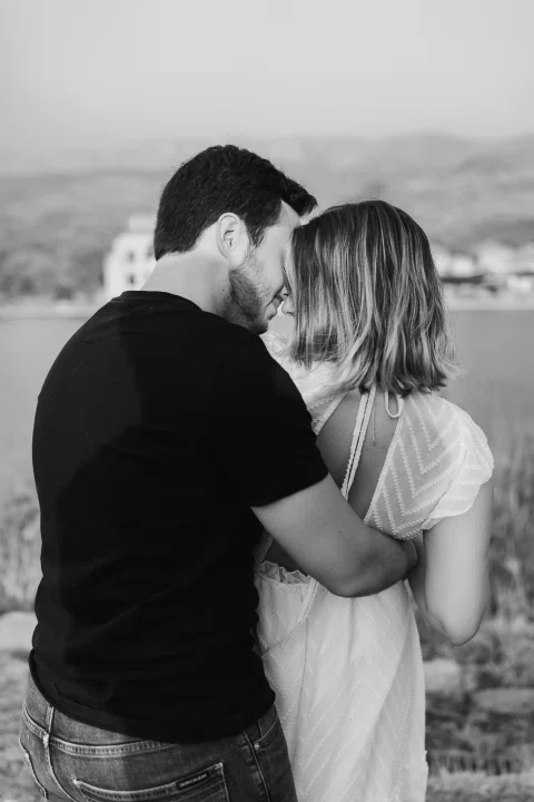 Hombre y mujer abrazados en un retrato cercano en blanco y negro, con agua y cerros al fondo.