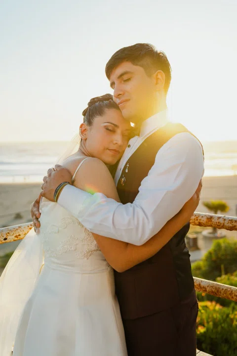 Hombre y mujer se abrazan a contraluz con la costa y el sol bajo al fondo.