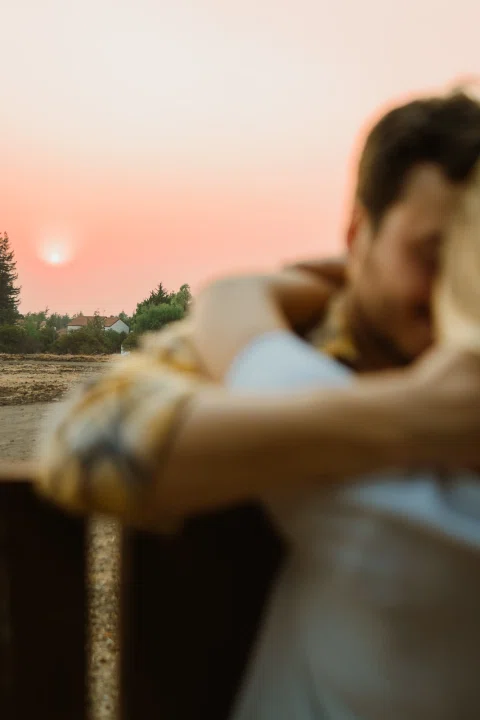 Hombre y mujer aparecen abrazados y desenfocados con luz de atardecer al fondo.