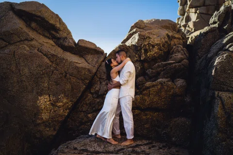 Hombre y mujer se abrazan entre grandes rocas junto a la costa.