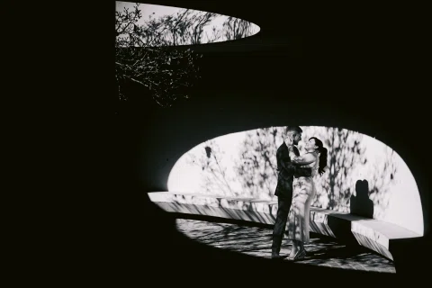 Hombre y mujer se abrazan dentro de una estructura curva iluminada, en una fotografía en blanco y negro.