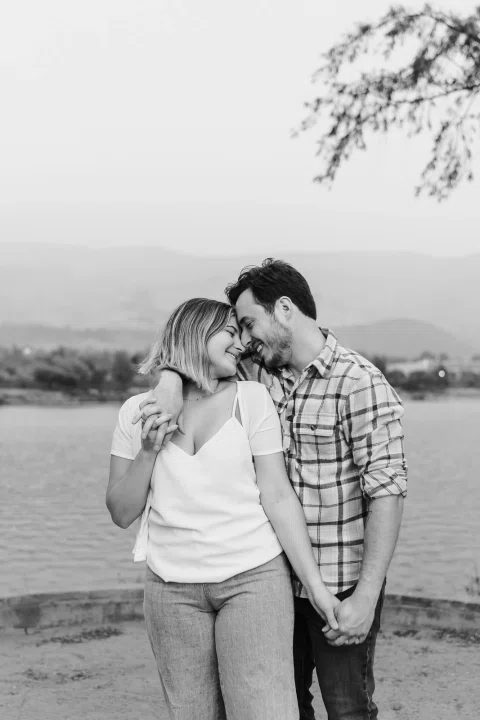 Hombre y mujer se abrazan con las frentes juntas frente a una laguna, en blanco y negro.