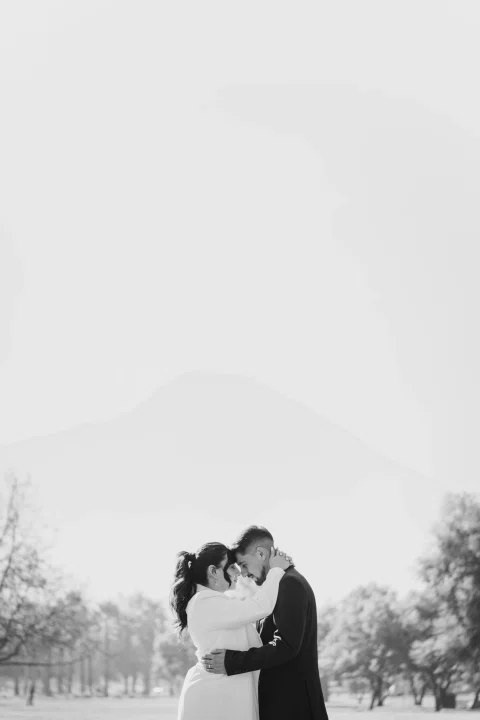 Hombre y mujer permanecen abrazados al aire libre con una montaña al fondo, en una fotografía en blanco y negro.