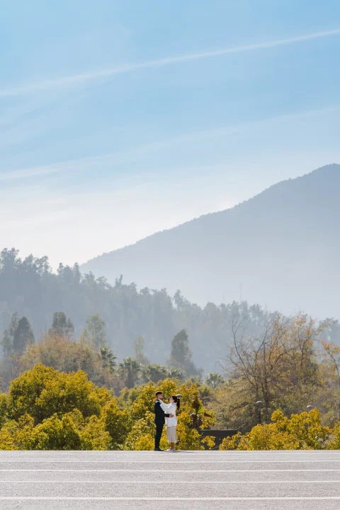 Hombre con traje azul y mujer con traje claro se abrazan a distancia en un paisaje de árboles y montañas.