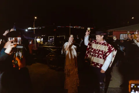 Hombre con poncho y sombrero y mujer con vestido largo levantan copas, rodeados por otras personas al aire libre durante la noche.