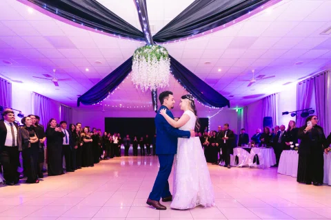 Hombre con traje azul y mujer con vestido blanco bailan abrazados en el centro del salón, rodeados por personas.