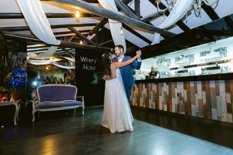 Hombre con traje azul y mujer con vestido blanco bailan abrazados frente a una barra.