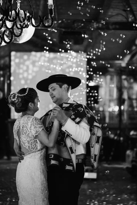 Hombre con poncho y sombrero y mujer con vestido blanco bailan juntos en una fotografía en blanco y negro, con otras personas al fondo.