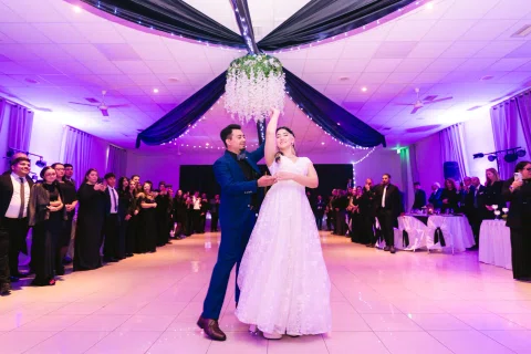 Hombre con traje azul y mujer con vestido blanco bailan mientras ella levanta un brazo bajo iluminación violeta.